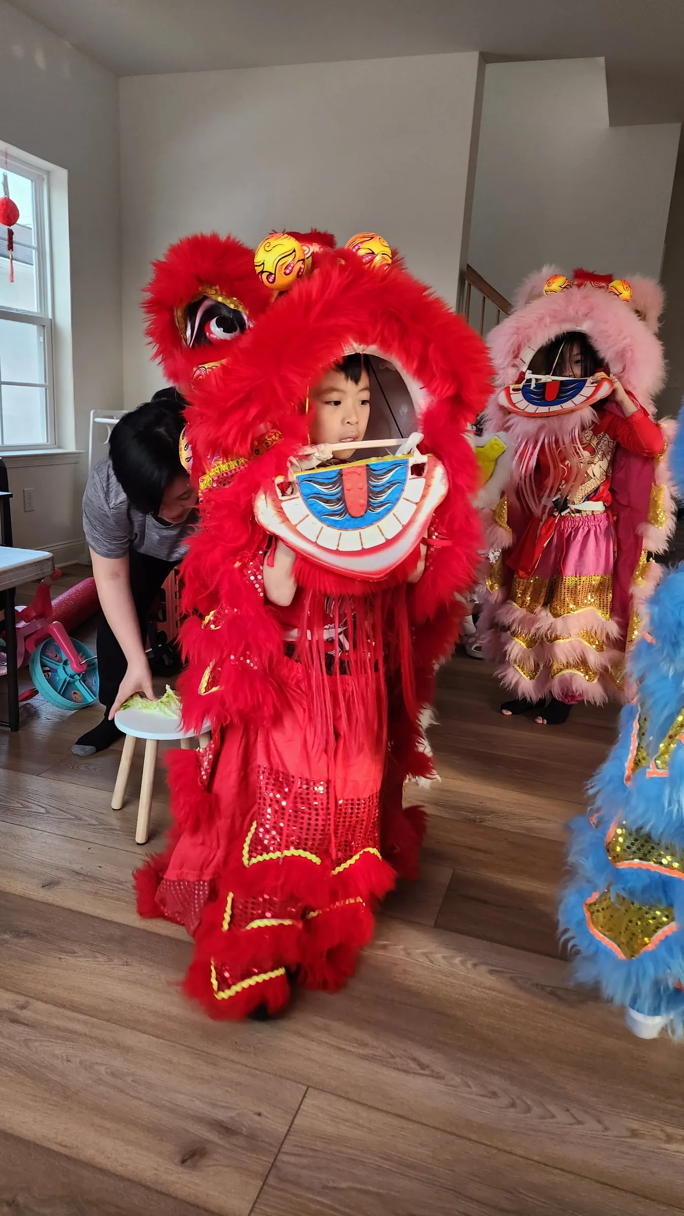 Young children trying on red and pink lion costumes indoors as an adult helps them